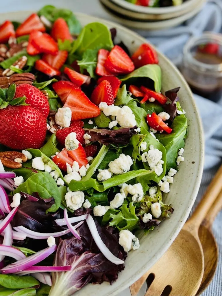 A large bowl heaping with green and purple salad mix, sliced bright red strawberries, brown pecans, thin slices of red onions, and white crumbles of dairy free bleu cheese.