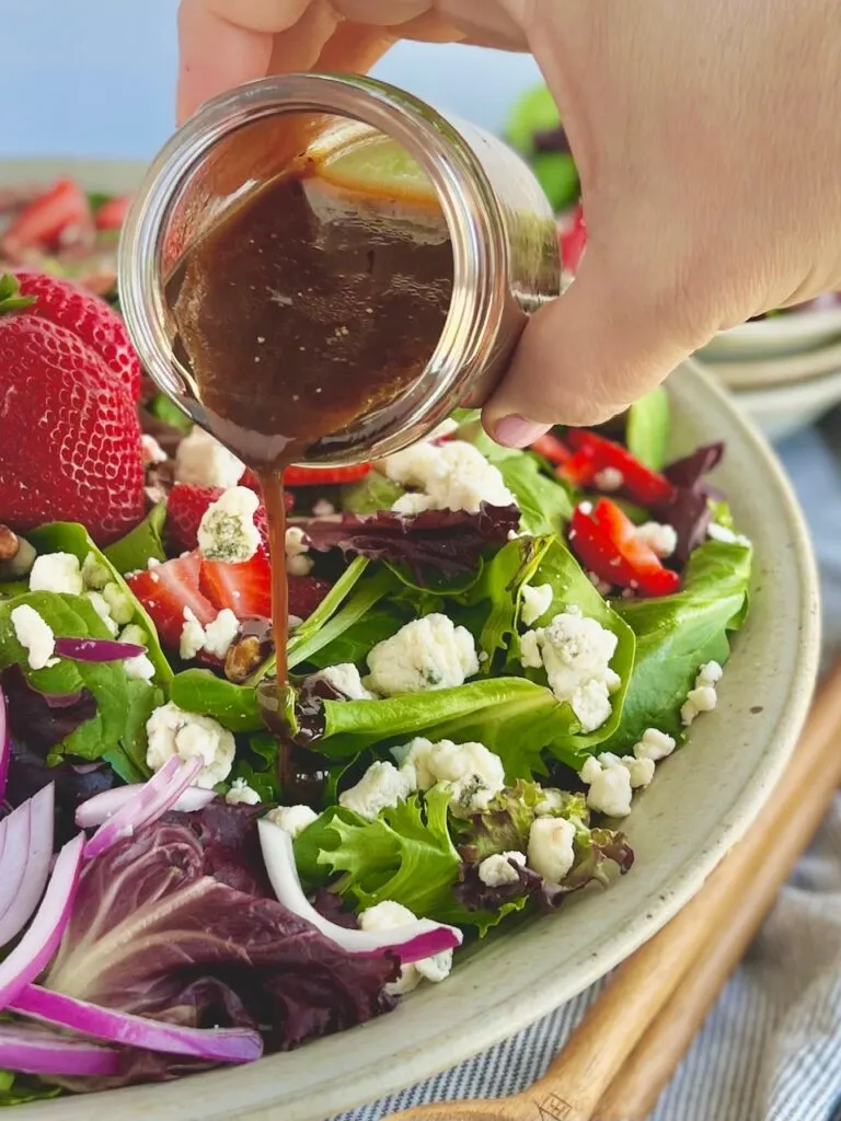 A hand pouring brown dressing into a large bowl heaping with green and purple salad mix, sliced bright red strawberries, brown pecans, thin slices of red onions, and white crumbles of dairy free bleu cheese.