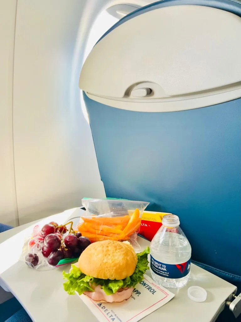 A sandwich, grapes, and cut bell peppers near an opened delta water sitting on a tray table of an airplane.