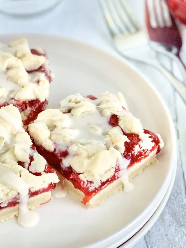 3 cherry bars on a white serving plate  with a bottom crust of a vanilla shortbread crust, topped with red cherry pie filling, another layer of white crumbly shortbread, and then it was drizzled with a white sweet glaze.