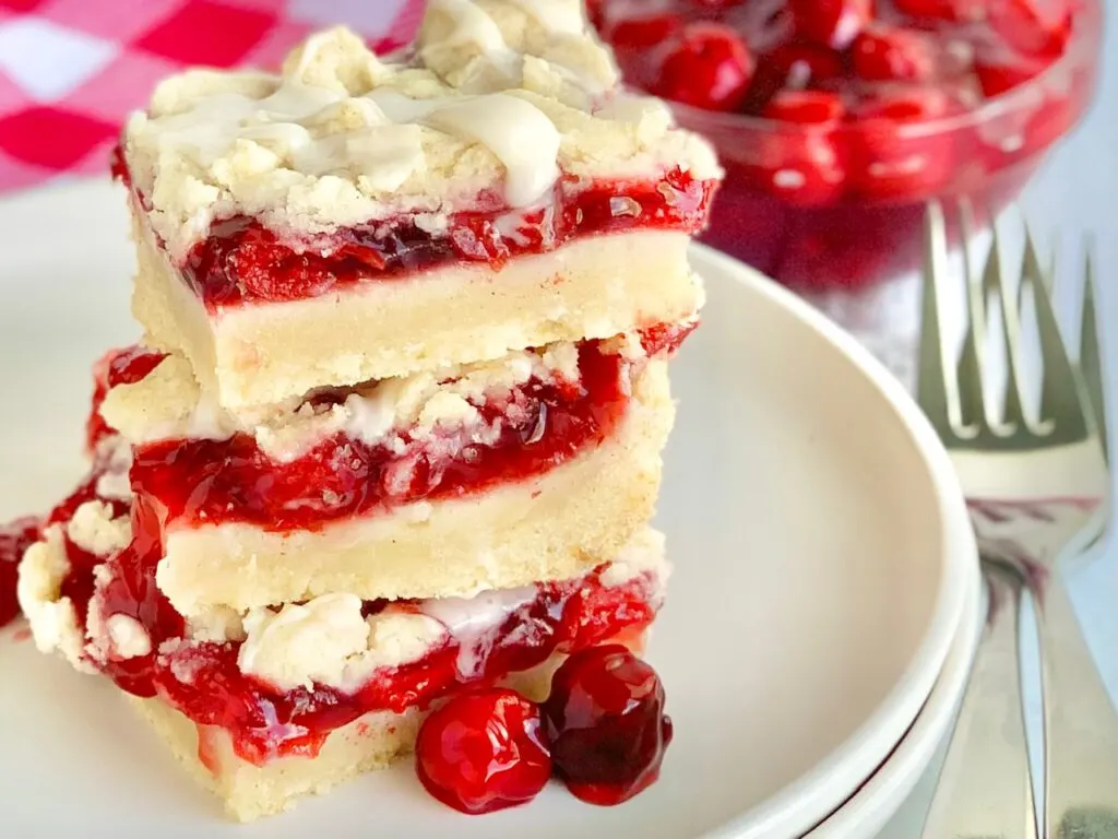 3 stacks of cherry bars with a bottom crust of a vanilla shortbread crust, topped with red cherry pie filling, another layer of white crumbly shortbread, and then it was drizzled with a white sweet glaze.