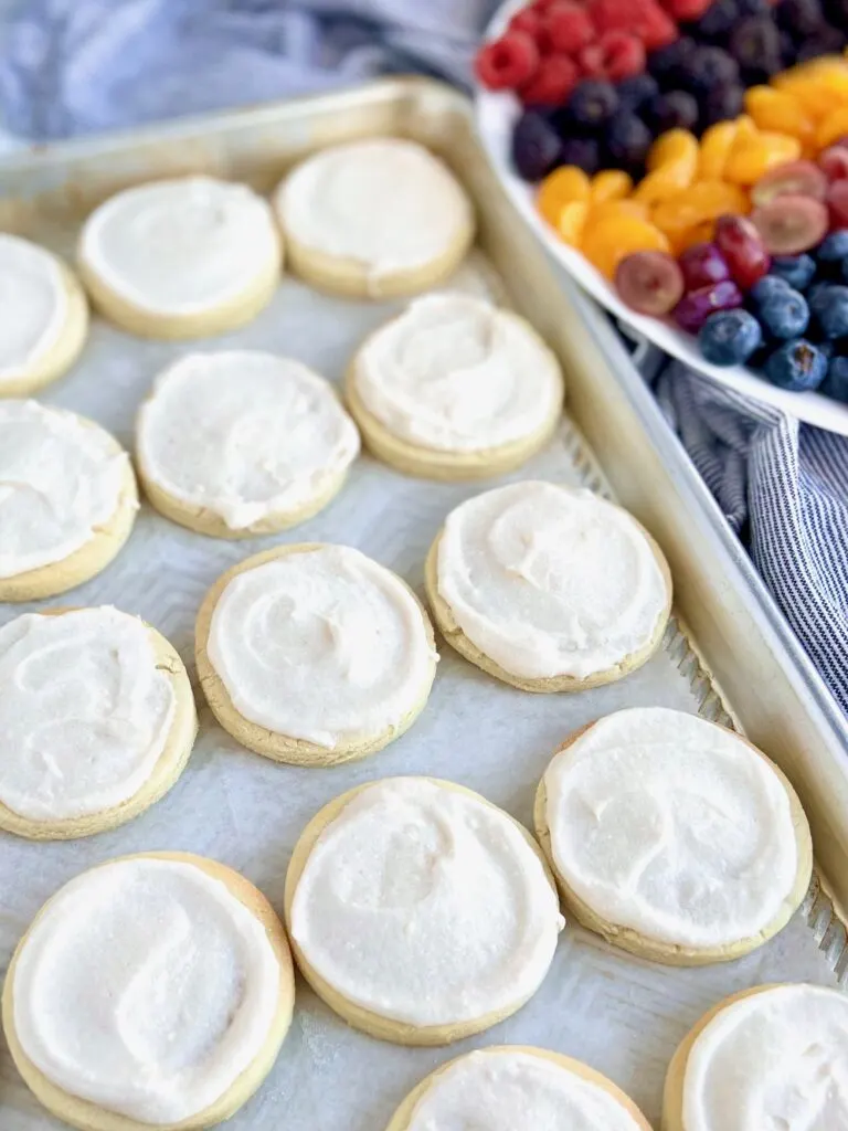 Several small round sugar cookies  with a white creamy frosting on top next to a plate of colorful fruit.