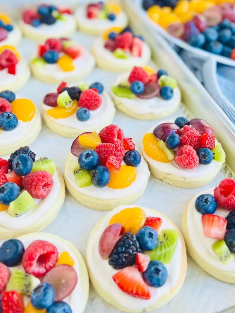 Several small round sugar cookies  with a white creamy frosting and a variety of fruit on the top such as blueberries, raspberries, sliced strawberries, sliced grapes, and mandarin oranges on a cookie sheet.