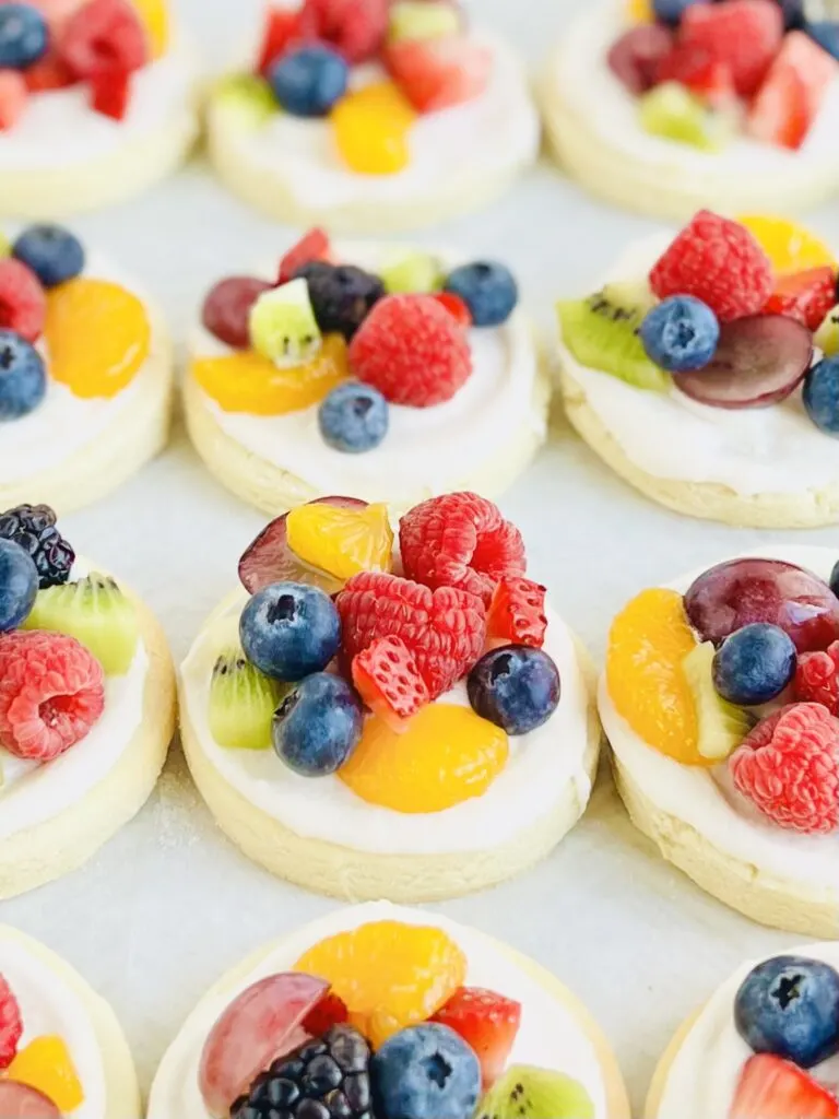 Several small round sugar cookies  with a white creamy frosting and a variety of fruit on the top such as blueberries, raspberries, sliced strawberries, sliced grapes, and mandarin oranges. 