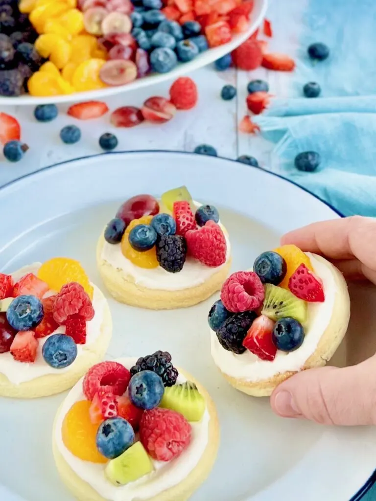 Four small round sugar cookies with a white creamy frosting and a variety of fruit on the top such as blueberries, raspberries, sliced strawberries, sliced grapes, and mandarin oranges with a hand reaching down to grab a cookie.