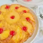 A round yellow brown cake with half pineapple slices around the the sides and a red Cherrie in the middle in the middle of the curved pineapple slice.