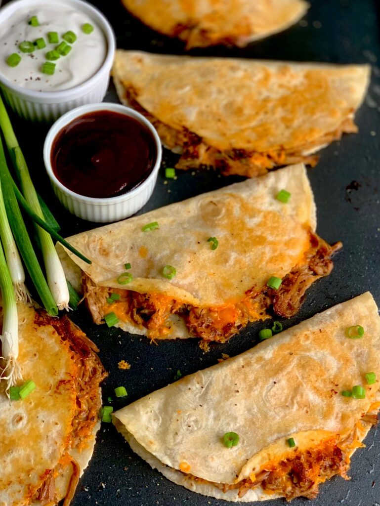 Three crispy looking tortillas with a brown pulled pork sticking out and yellow melted cheese on a black griddle next to a small bowl of BBQ sauce and green onions.