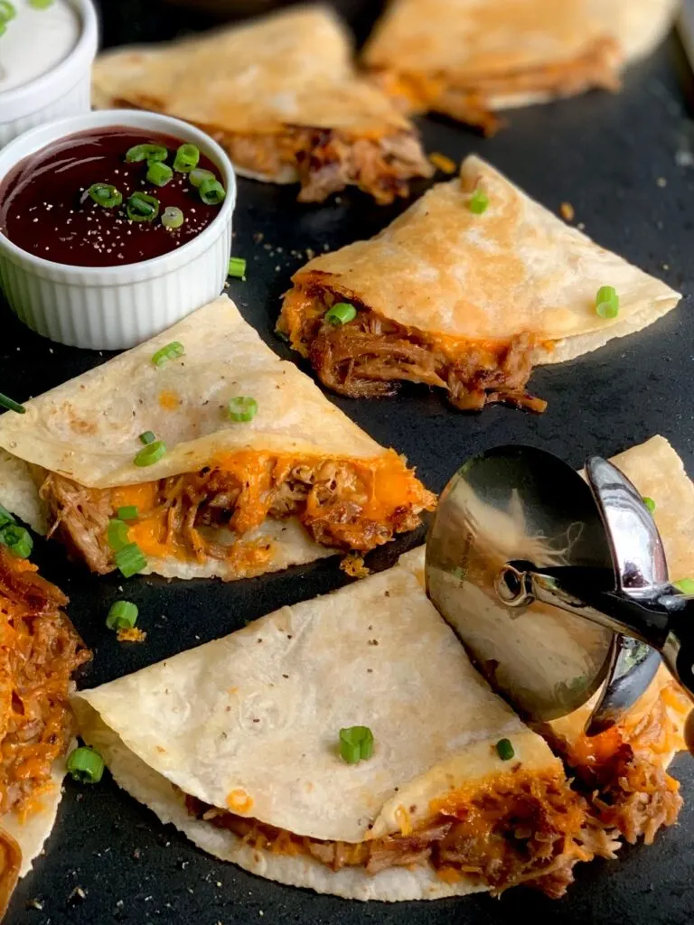 Several crispy looking tortillas with a brown pulled pork sticking out and yellow melted cheese on a black griddle next to a small bowl of BBQ sauce.