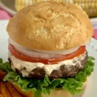 A hamburger loaded with green lettuce, a thick juicy looking hamburger topped with an orange sauce, a thin cut tomato, and sliced of white onion all on a white plate.