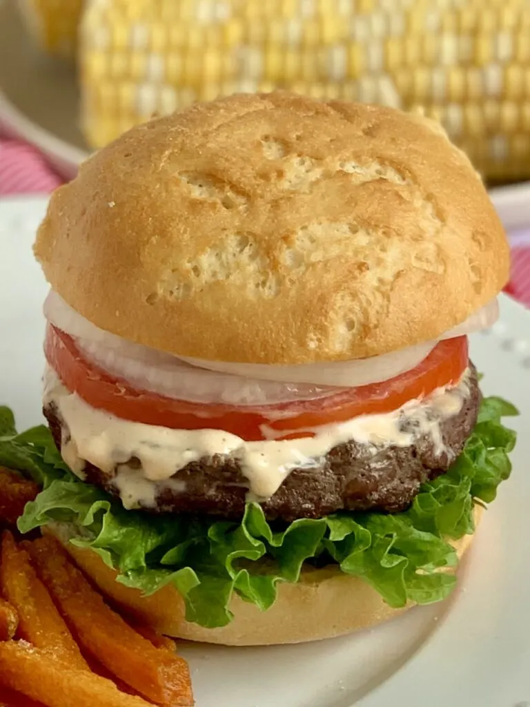 A hamburger with green lettuce on the bottom, topped by a juicy looking hamburger, a thick orange sauce, thin sliced tomato and white onions next to sweet potato fries on a white plate.