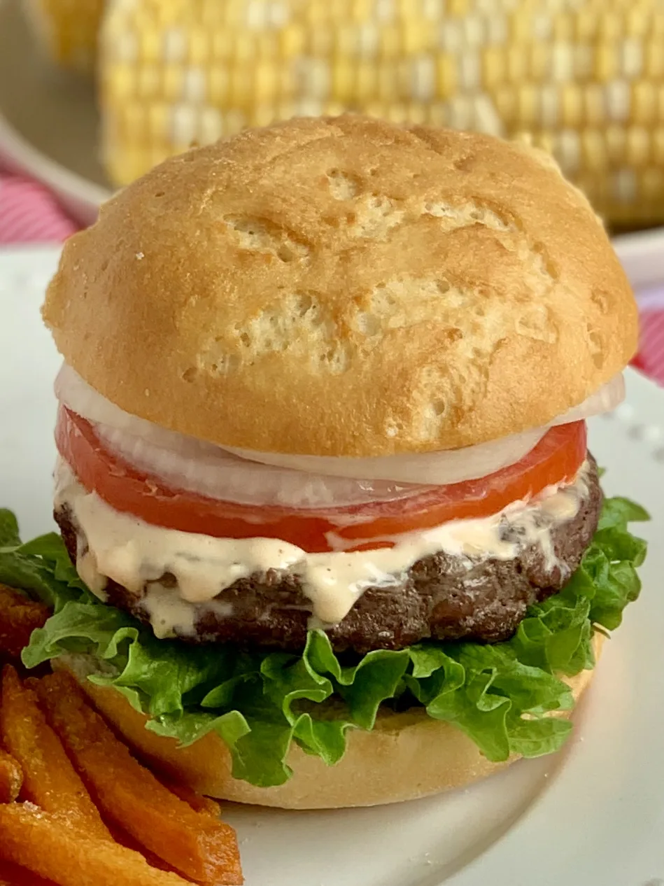 A hamburger loaded with green lettuce, a thick juicy looking hamburger topped with an orange sauce, a thin cut tomato, and sliced of white onion all on a white plate.