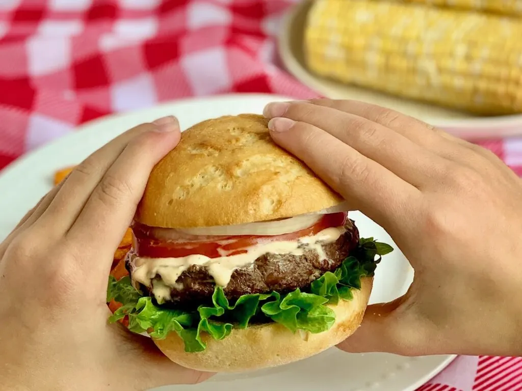 Hands holding a hamburger with green lettuce on the bottom, topped by a juicy looking hamburger, a thick orange sauce, thin sliced tomato and white onions.