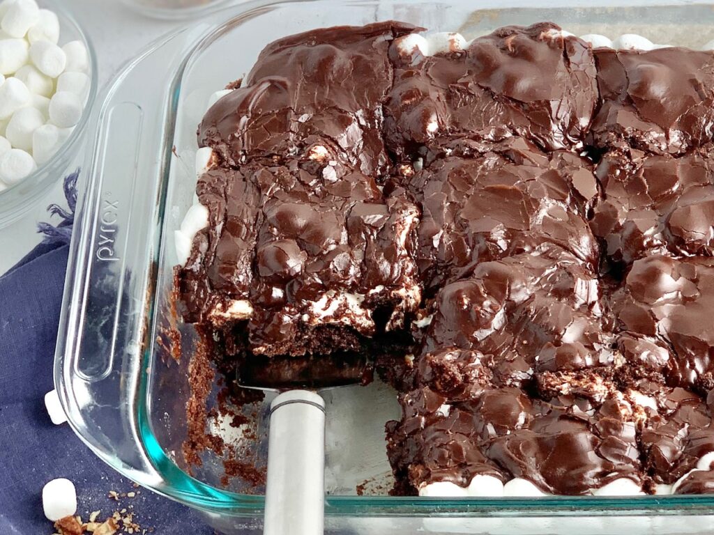 A glass baking dish filled with a dark brown chocolate cake followed by a white layer of mini marshmallows and topped with dark brown chocolate frosting. The bars have been cut into squares and some are missing from the pan. A spatula has lifted a slice out of the pan.