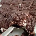 A glass baking dish filled with a dark brown chocolate cake followed by a white layer of mini marshmallows and topped with dark brown chocolate frosting. The bars have been cut into squares and some are missing from the pan.m,
