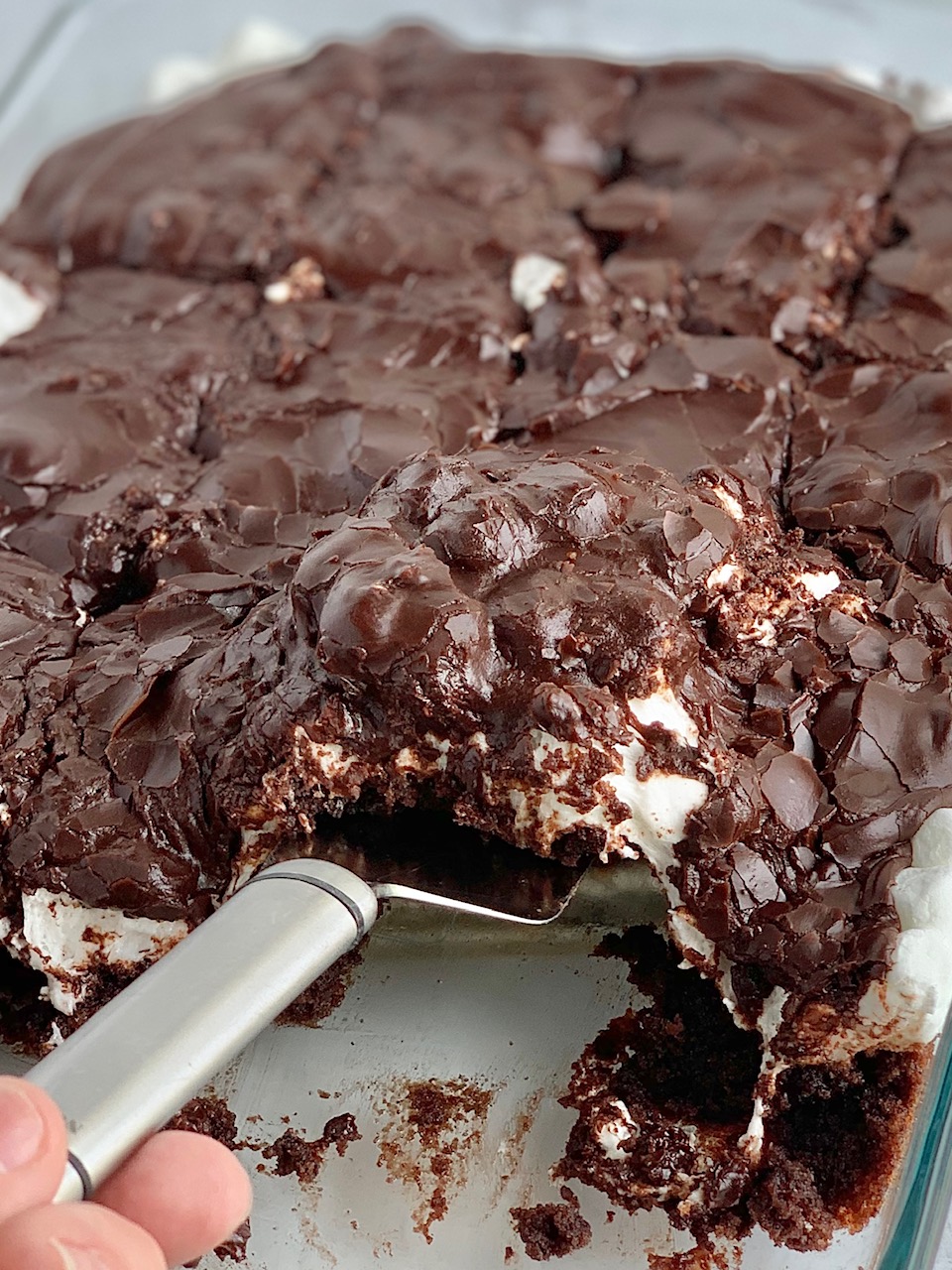 A glass baking dish filled with a dark brown chocolate cake followed by a white layer of mini marshmallows and topped with dark brown chocolate frosting. The bars have been cut into squares and some are missing from the pan.m,