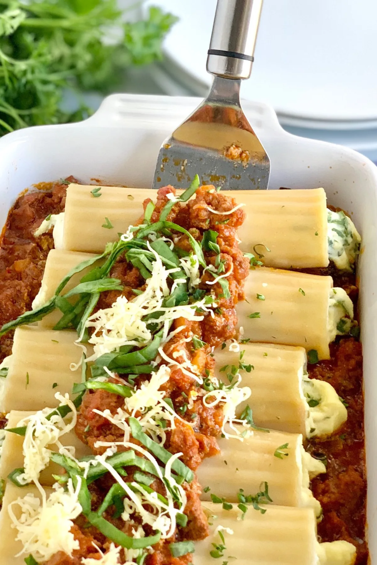 A large white baking dish filled with ground beef and marinara sauce then topped with round pasta shells filled with spinach, and a ricotta mixture. Then that's topped with more ground beef marinara sauce, dairy free cheese shreds, spinach, and fresh parsley with a serving spatula lifting up two of the manicotti out of the pan.