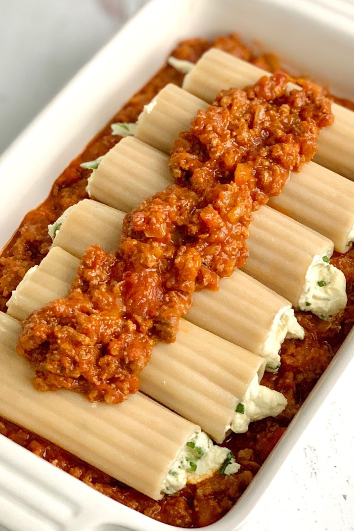 A large white baking dish filled with ground beef and marinara sauce then topped with round pasta shells filled with spinach, and a ricotta mixture. Then that's topped with more ground beef marinara sauce, 
