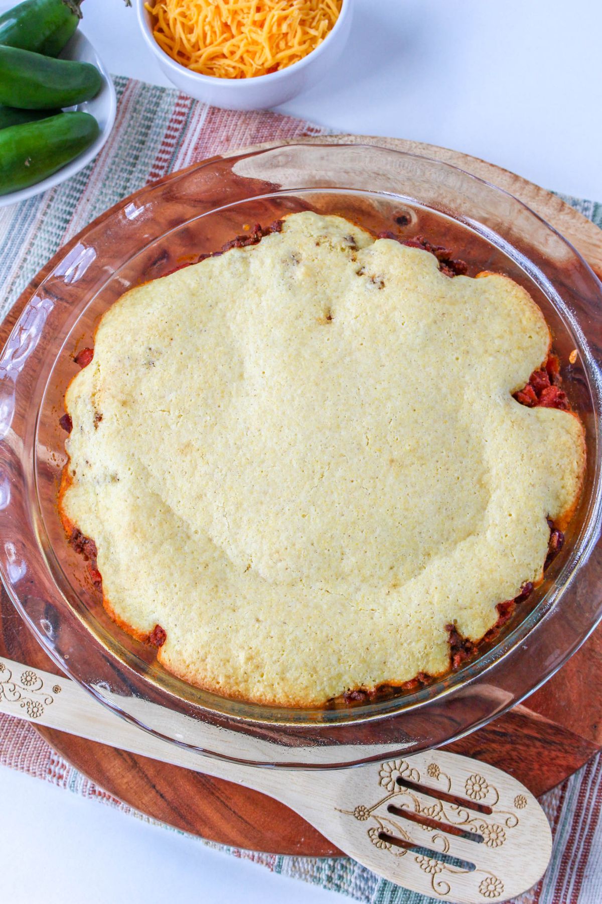 A serving dish with the wooden spoon next to a chili pie with cornbread topping.