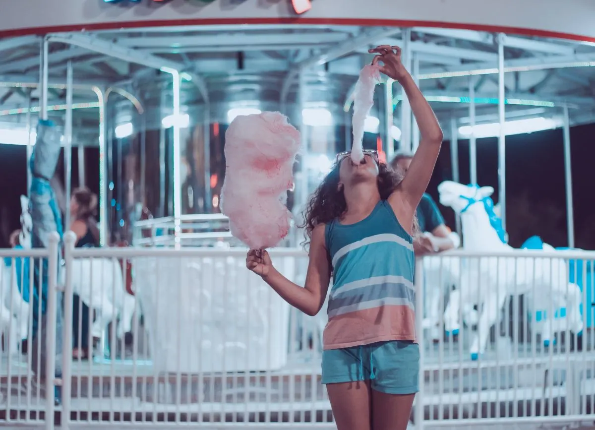 A girl putting cotton candy in her mouth standing in front of a carousel.